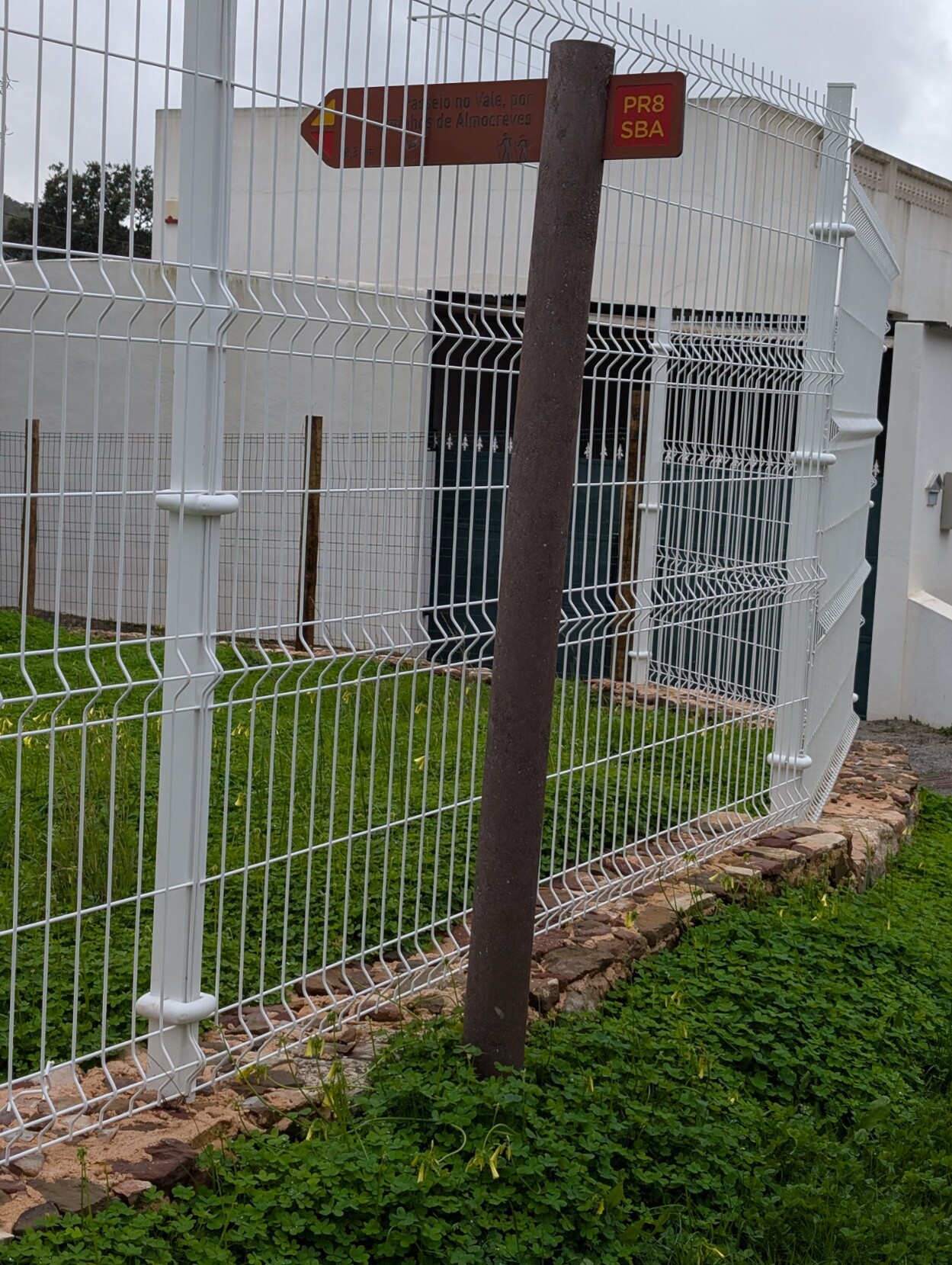 A footpath finger post where a white metal wire fence has been constructed but to make way for the finger of the post the metal fence has been left incomplete at that point.